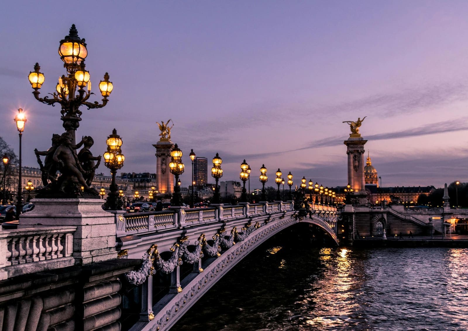 Gilded lamps and statues along an ornate stone bridge over a river at dusk in Paris