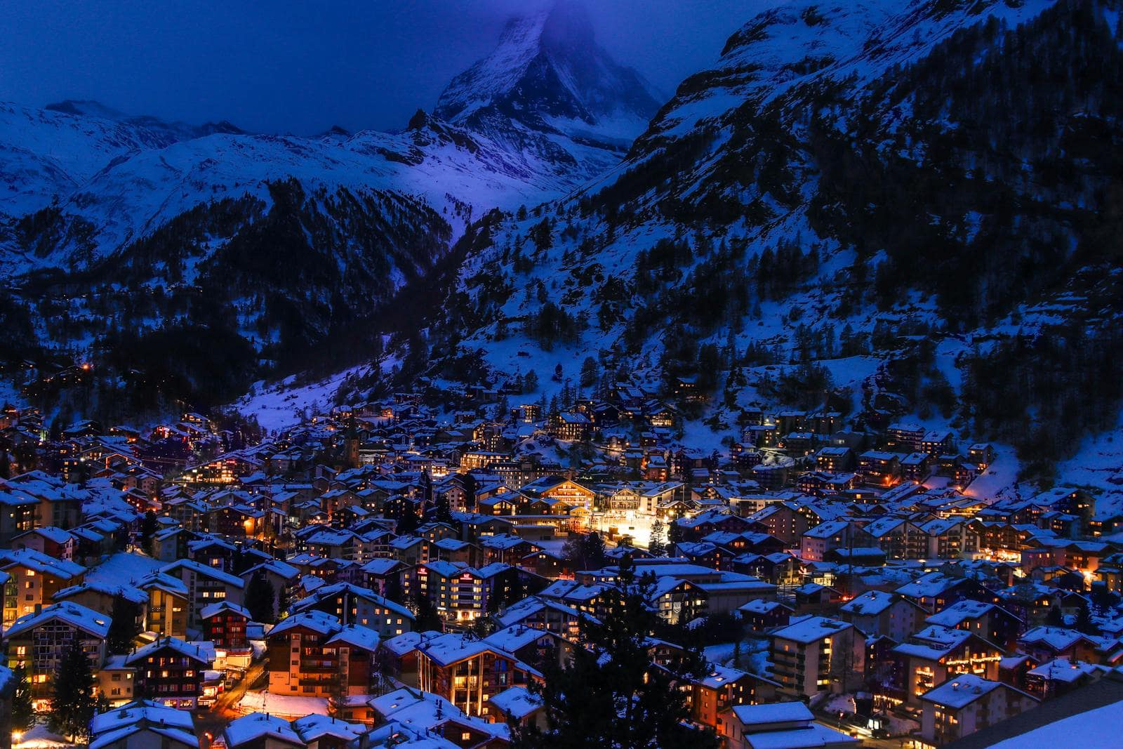 A snow-covered alpine town glowing with window lights at blue hour beneath a sharp peak
