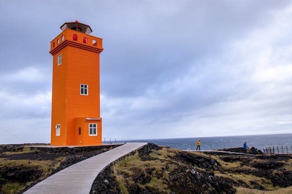 A bright orange lighthouse at the end of a boardwalk on a rocky coast under grey cloud
