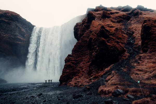 Three tiny figures dwarfed by a wide waterfall pouring over a dark cliff